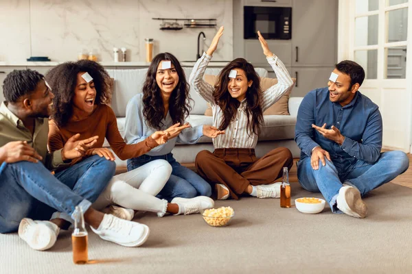 Cheerful multicultural students friends playing guess who game, enjoying beer and popcorn, sitting on floor in modern living room, chilling together at home on weekend, having cards on foreheads