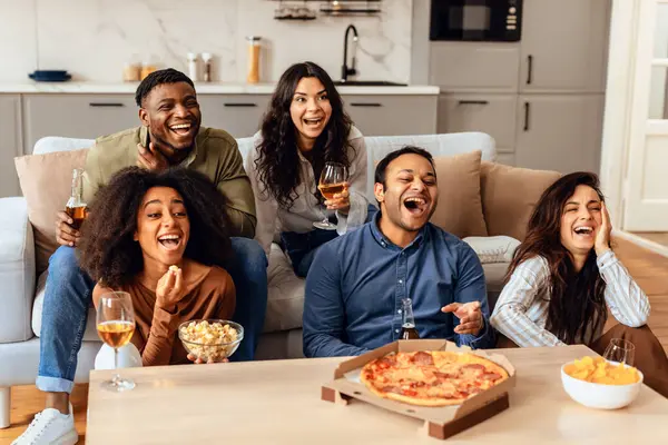 Happy group of diverse friends watching comedy film at home, eating popcorn and pizza enjoying funny day together, relaxing on sofa with drinks and junk food. Friendship concept