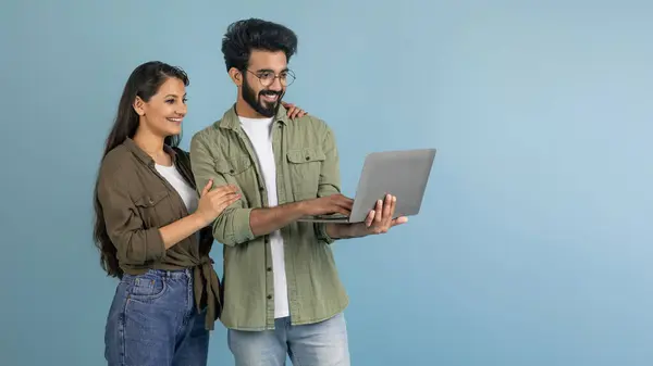 陽気なミレニアルインドカップルは 一緒にラップトップを使用してカジュアルな服を着て コンピュータ画面と笑顔 青いスタジオの背景 コピースペースを持つパノラマを見ています オンラインショッピング — ストック写真
