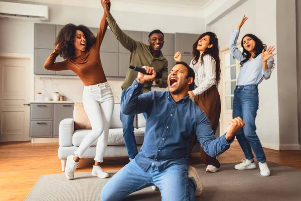 Party with friends. Indian guy with microphone singing karaoke song while diverse friends group dancing, having fun together during students gathering on weekend at home interior
