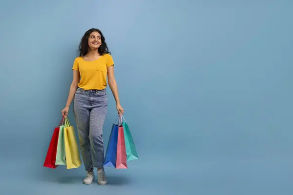 Shopping, sale season, black friday deal. Happy beautiful young indian woman wearing casual clothing with colorful paper bags purchases posing on blue background, looking at copy space, full length