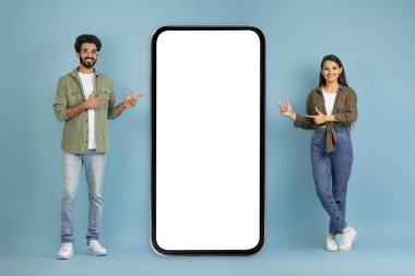 İki kişi için harika bir teklif. Neşeli genç Hintli çift beyaz ekranlı akıllı telefondan poz veriyor, en yeni mobil uygulamaları gösteriyor, model, kopyalama alanı, mavi arkaplan
