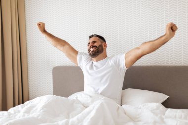 Avrupalı sakallı bir adam modern yatak odasında uyanıyor ve kollarını mutlu bir gülümsemeyle açıyor, beyaz çarşaflarla yatakta oturuyor. Ev konseptinde rahat bir sabah rutini.