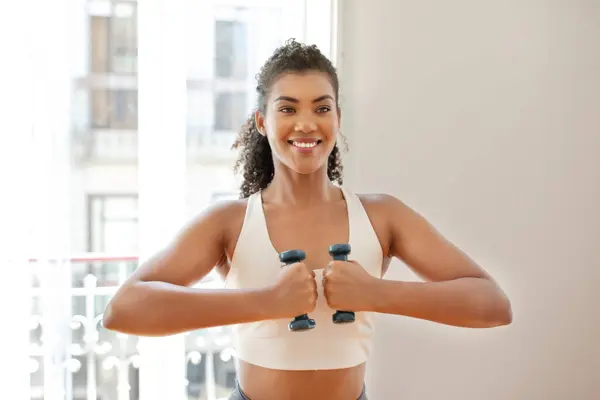 Happy fit woman holds dumbbells while confidently works out with focus on bodybuilding at home, smiling to camera exercising for arms muscles strength. Morning training routine, fitness workout
