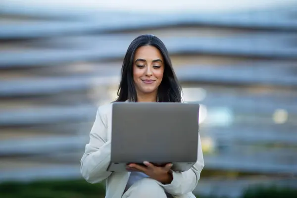 Hispanic business lady browsing websites on her laptop sitting outside city office center, smiling as she works online on computer gadget, working as freelancer in urban area