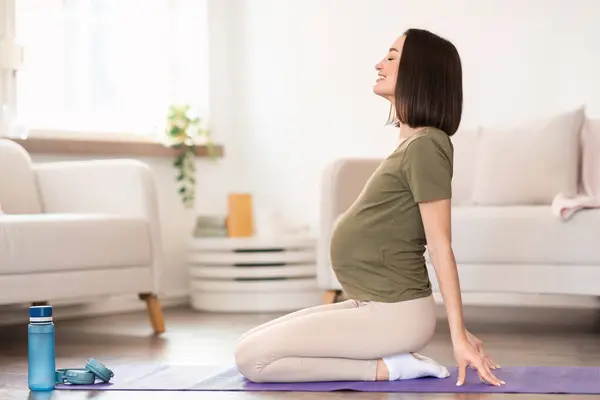 Pregnant lady doing back bend sitting on mat during yoga class, exercising at cozy home. Sporty woman expecting baby, enjoying stretching workout and healthy lifestyle. Side view shot, copy space