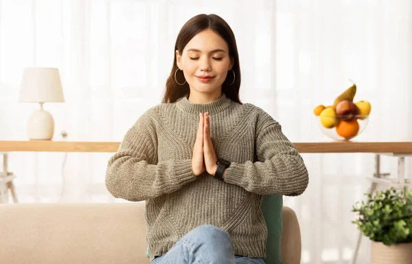 Calm young brunette woman sitting on couch with closed eyes and smile on her face, concentrating or meditating, praying in the morning, holding hands in namaste gesture, home interior