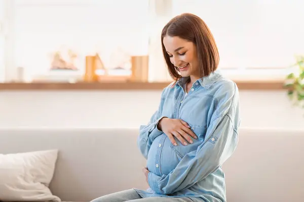 Szczęśliwa Oczekująca Kobieta Ciąży Siedzi Kanapie Salonie Dotykając Brzucha Uśmiechając — Zdjęcie stockowe