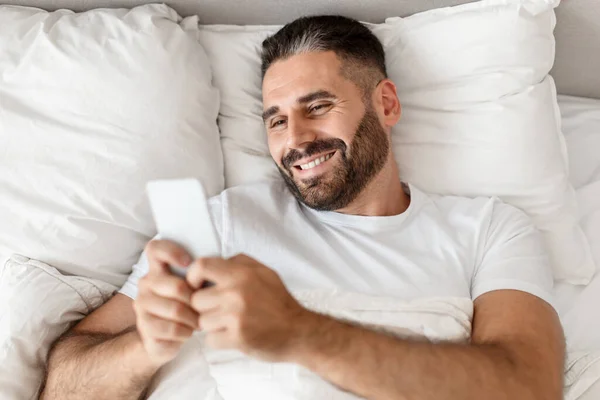 Top view of middle aged European man casually scrolling through his phone in bed, lying and resting in the morning, browsing emails and social media in bedroom. Modern technology lifestyle