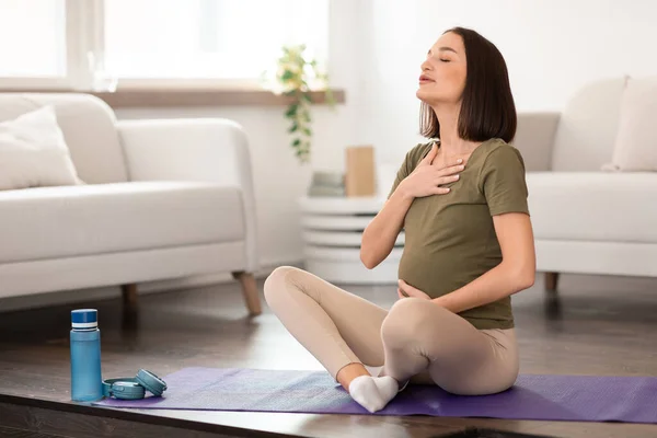 Pregnant woman practices prenatal yoga, pressing hand against chest while focusing on deep breathing exercises in modern domestic living room. Sport, pregnancy and wellbeing