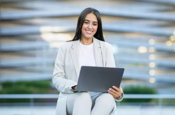 Happy Arabic businesswoman working on laptop sitting in front of modern office building in city, showcasing her expertise in software and digital technology. Successful programmer concept