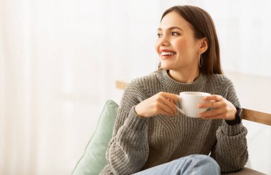 Huzurlu genç esmer kadın kanepede oturuyor, evde sıcak kahve içiyor, fotokopi odasına bakıyor ve gülümsüyor. Neşeli bayan oturma odasında sıcak bir içkiyle tembelliğin tadını çıkarıyor.