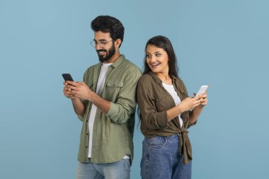 Elinde akıllı telefonuyla gülümseyen Hintli genç bir kadın cep telefonu kullanarak erkek arkadaşını gözetliyor, mavi stüdyo arka planında izole bir şekilde kocasına makaralar yolluyor.