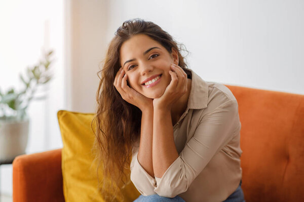 Beautiful young woman with wavy hair smiling and resting head on her hands while relaxing on comfortable couch in living room, happy female posing at camera while resting at home, free space