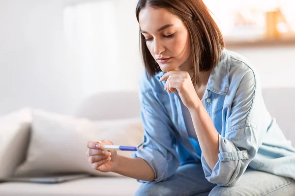 Unhappy young lady holds negative pregnancy test with sad and anxious expression, sitting in modern living room at home. Unwanted pregnancy or infertility health issue concept