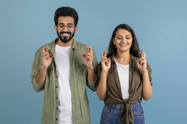 Excited millennial indian couple man and woman wearing casual clothing holding crossed fingers, posing with closed eyes, make wish, cherish hope, isolated on blue studio background