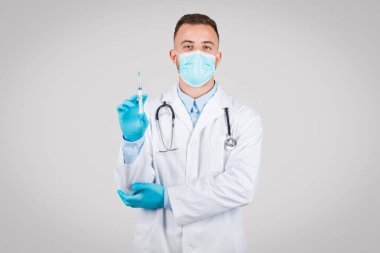 Koruyucu maskeli ve mavi eldivenli erkek doktor, aşıya veya tedaviye hazırlık durumunu temsil eden tıbbi bir şırınga tutuyor.