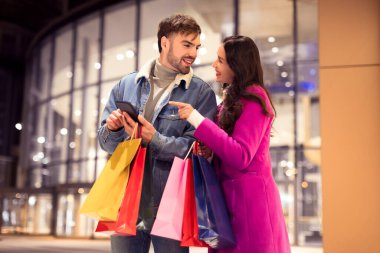 Genç çift kış şenliği sırasında online satış alışverişi için telefonları kontrol ediyor, kış şehrinde akşamları alışveriş torbalarıyla modern alışveriş merkezinin önünde duruyorlar. Noel ve Sevgililer Günü indirimi