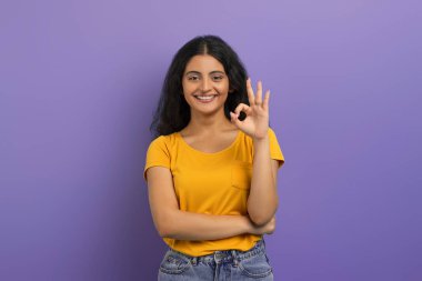 Neşeli, güzel, günlük kıyafetler giyen, iyi bir jest yapan, kameraya gülümseyen, mor stüdyo arka planında yalnız poz veren güzel Hintli bir kadın. Her şey yolunda.