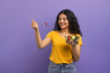 Çekici gülümseyen kıvırcık saçlı, günlük giysiler giyen, mor stüdyo arka planında izole edilmiş poz veren sağlıklı yemek yiyen, taze sebze salatası yiyen genç Hintli kadın. Diyet, beslenme, kilo kaybı