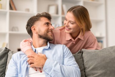 Millennial karısı kocasına sevgiyle ve sıcakkanlılıkla gülümseyip evdeki kanepede poz veriyor. Genç Avrupalı eşler sevgi dolu bir kucaklaşmayı paylaşıyorlar. Mutlu evlilik anları konsepti