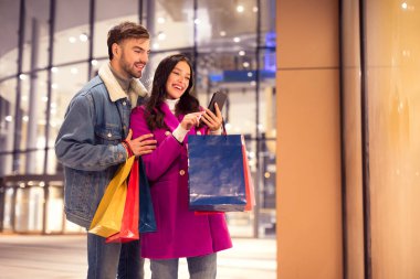 Milenyum Avrupalı çiftler kış akşamı modern alışveriş merkezlerinin yakınında telefonla alışveriş yapıyorlar, alışveriş torbalarıyla duruyorlar, Noel ve yeni yıl hediyeleri alıyorlar. Boş Boşluk