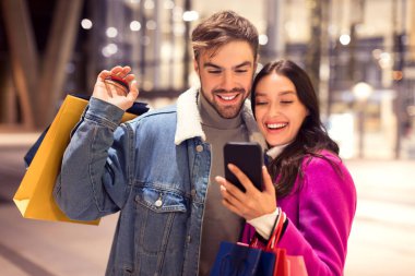 Kışın giyinmiş genç bir adam ve kadın cep telefonu uygulamasını online alışveriş, web sörfü satışları ve anlaşmaları için kullanıyor, Noel 'de ya da yeni yılda alışveriş torbalarıyla ya da dışarıda bekliyor.