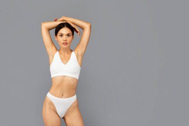 Sakin, beyaz iç çamaşırlı Asyalı genç bir kadın gri arka planda, elleri başının üstünde, kameraya bakarak koltukaltlarını gösteriyor. Fitness, kuru cilt, tazelik