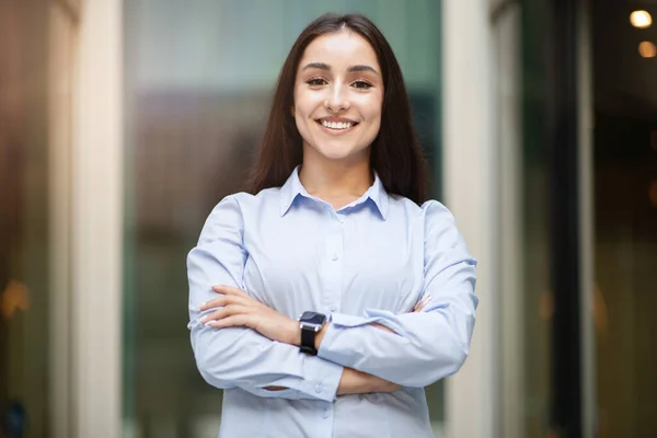 Confident arab young professional woman with long hair and blue shirt, standing with arms crossed, smiling at camera, with smartwatch. Professional and modern work, career
