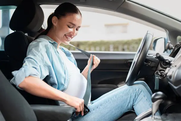 Young pregnant woman in her car adjusting seat belt for comfort and safety, posing in drivers seat, preparing for ride through city. Journey of motherhood concept. Side view