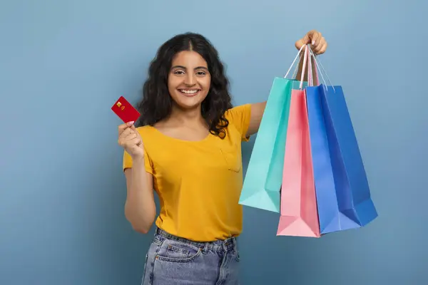 Sales And Discounts Concept. Smiling excited young indian woman holding credit card and shopping bags, got cashback, looking at camera, standing isolated over blue studio background