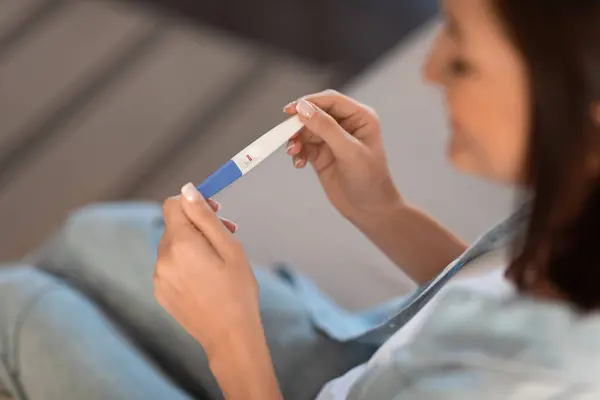Smiling pregnant lady holds positive pregnancy test sitting indoor, high angle view, cropped shot. Fertility and childbirth, motherhood concept. Selective focus on medical test