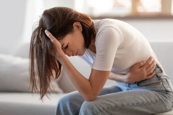 Young lady touching her stomach and head in distress, suffering from headache and stomachache, having symptoms of PMS and gastrointestinal discomfort, sitting in living room at home