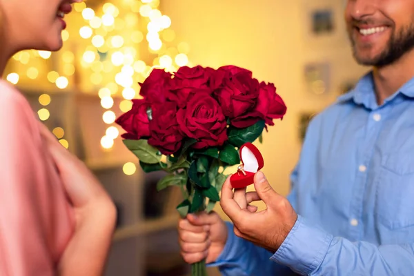 暖かく照らされた部屋で 男性は結婚提案で彼のパートナーを驚かせ 赤いバラの花束と一緒に輝く婚約指輪を提示します — ストック写真