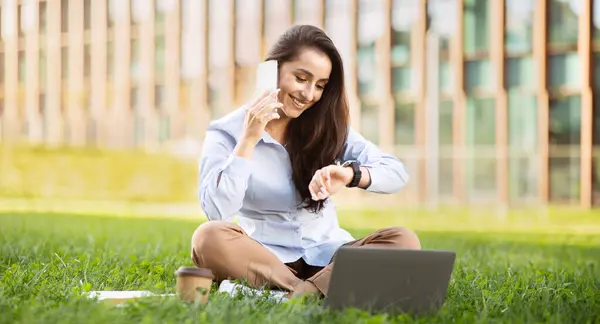 Multitasking happy european young woman sits on grass with laptop, talking on phone, checking watch, outdoor, panorama. Efficient time management in relaxed setting, planning project