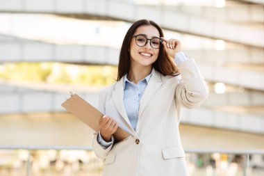 Gözlüklü neşeli genç bir iş kadını elinde bir pano tutarken çerçevesini ayarlıyor. Havadar, modern bir ofis ortamında kendine güven ve profesyonellik yayıyor. Profesyonellik ve güven