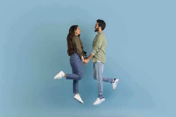 Happy eastern lovers millennial man and woman wearing casual clothing holding hands and jumping up in the air, celebrating success, isolated on blue studio background, side view