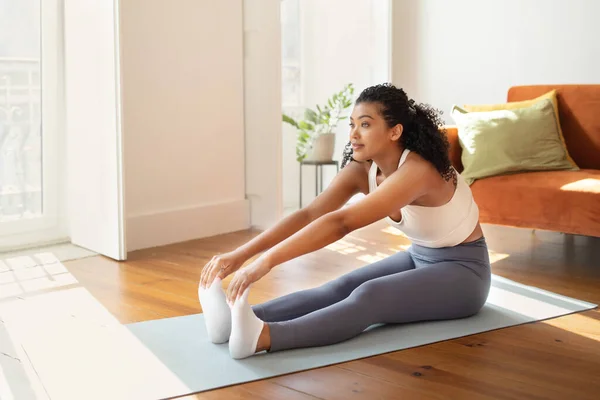 Fit young woman in sportswear stretching on yoga mat, performing seated toe touch exercises in her living room, embodying active and healthy lifestyle in morning fitness routine