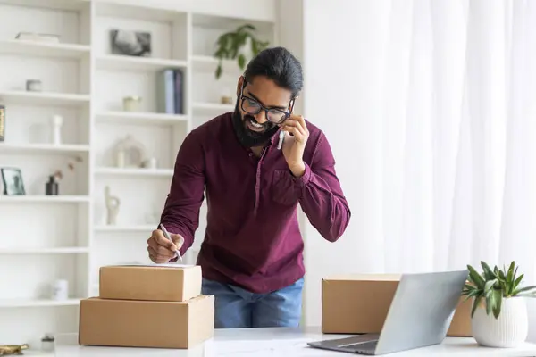 Indian Man Checking Orders While Working At Home Office, Smiling Young Eastern Male Standing Near Desk With Packed Boxes, Speaking With Clients By Cellphone, Enjoying Managing Small Business