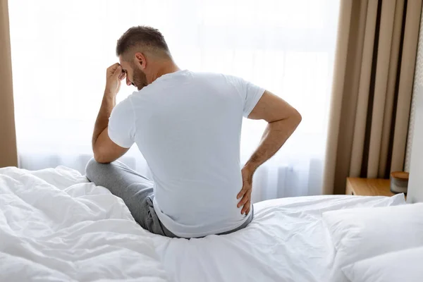Unrecognizable European man touching lower back and head suffering from painful backache, sitting on the edge of bed at home. Guy struggles with spinal discs problems. Rear view