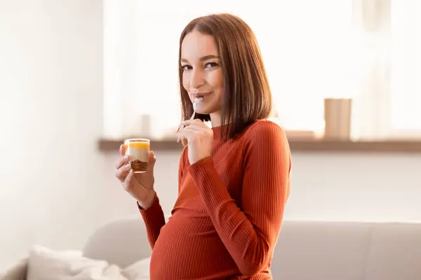 年轻的孕妇喜欢吃美味的小吃 吃甜食 对着镜头笑 平衡怀孕期间健康饮食的需要和即将分娩的渴望 站在室内 — 图库照片