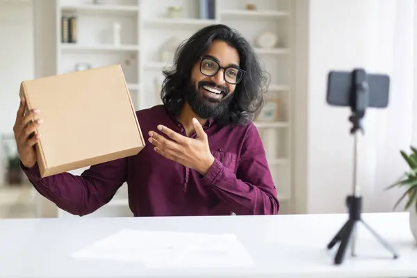 Video Review. Young Indian Man Demonstrating Cardboard Box At Smartphone Camera, Smiling Eastern Male Recording Content For His Vlog, Blogger Guy Making Unboxing While Sitting At Desk At Home