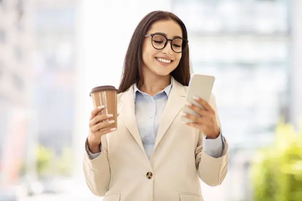 Content businesswoman in a stylish beige jacket holds a coffee cup and smartphone, engaged in reading the screen outdoors. Modern multitasking and technology use on the go