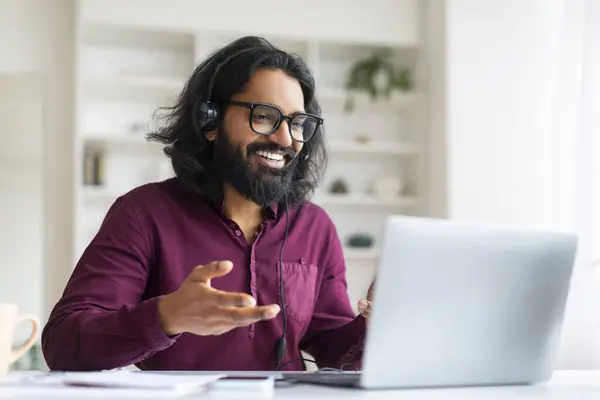 Cheerful indian freelancer wearing glasses and headset making video conference on his laptop while sitting at desk in home office, happy handsome eastern male enjoying online communication