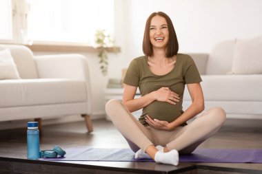 Neşeli genç hamile bayan yoga minderinde oturur, kameraya gülümserken sevinçten karnına sarılır, hamilelik boyunca sportif ve sağlıklı yaşam tarzının tadını çıkarır. Doğum öncesi yoga ve meditasyon konsepti