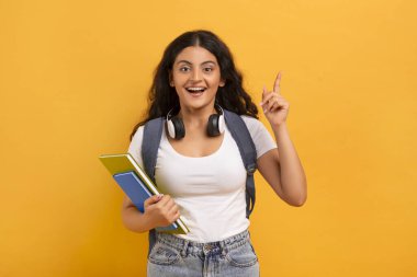 Sırt çantası ve çalışma kitaplarıyla heyecanlı bir genç doğu öğrencisi. Parmağını kaldırıp kameraya bakıyor. Harika bir yaratıcı fikri, eğitim projesi, sarı stüdyo geçmişi var.