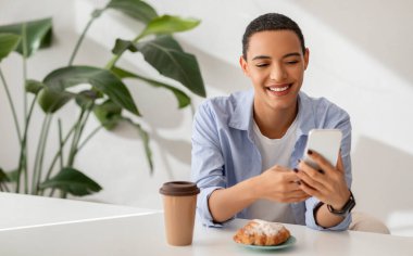 Gündelik kahve molası veren mutlu genç bir kadın, akıllı telefonuyla mesaj atıyor, yanında taze bir pasta, arka planında da kapalı alanda bitkiler var. Kahve molası ve dijital bağlantı.