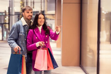 Sevgililer Günü hediyeleri için gülümseyen ve heyecanlı genç çiftler mağazaları, ellerinde hediye paketleri, şehir merkezinin önünde durup kış akşamları mağazanın camını işaret ediyorlar. Boşluk alanı