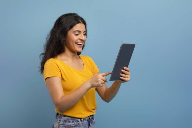 Kot pantolon ve sarı tişört giyen mutlu Hintli bayan. Alet dijital tableti kullanıyor, arkadaşlarıyla sohbet ediyor, web sörfü yapıyor, sosyal medyada kayıyor, mavi arka planda izole edilmiş, uzay kopyaları yapıyor.