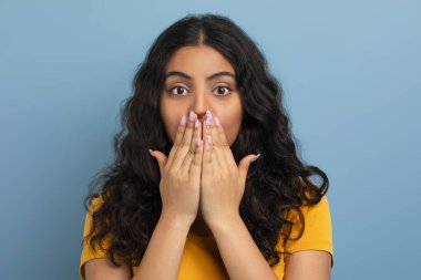 Sarı tişört giyen, şaşırtıcı kıvırcık, genç, doğulu bir kız öğrencinin elleri ile ağzını kapatması, mavi stüdyo arka planında şok etkisi yaratıyor. İnsan duyguları kavramı